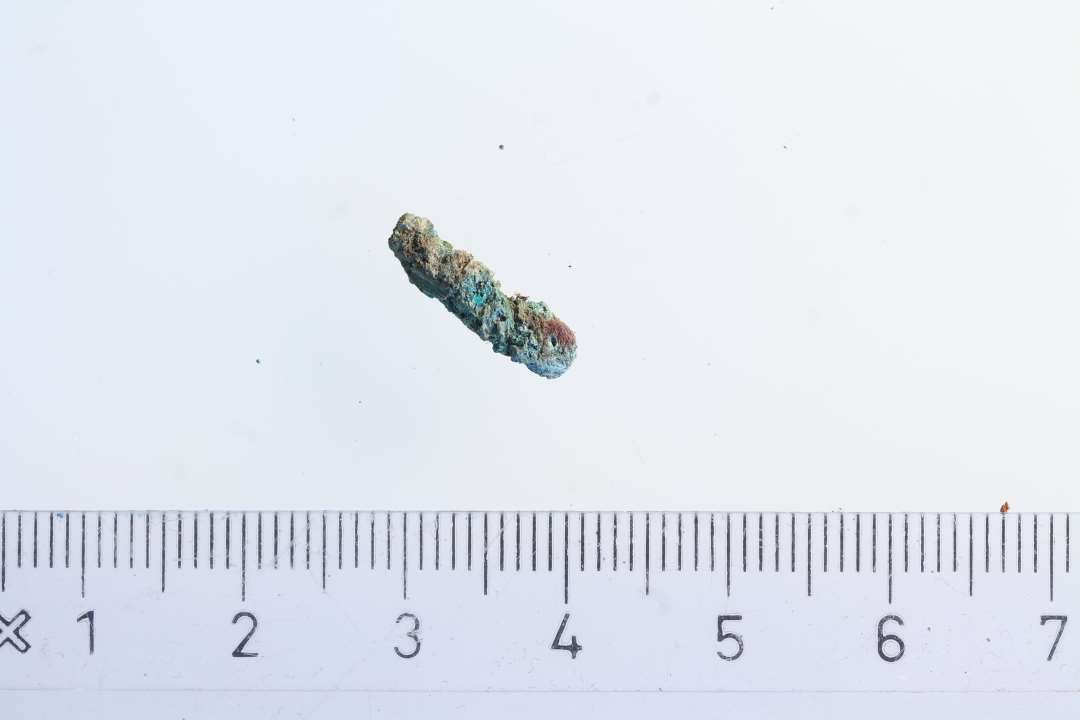 Lille stykke bronze p� ca. 1,2 cm. En mulig torn fra et dragtsp�nde, med et meget lille stykke r�d/lilla tekstil, der synes at v�re p� begge sider.  