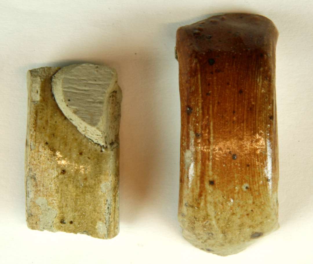 To fragmenter af b�jleformede hanke antagelig fra kander af gr�ligt finslemmet stent�j. Hankene har flad oval tv�rsnitsform, den ene med flad hulkehl p� ydersiden. Begge fragmenter har uj�vn r�dbrun bemaling hvorover klar slatglasur.