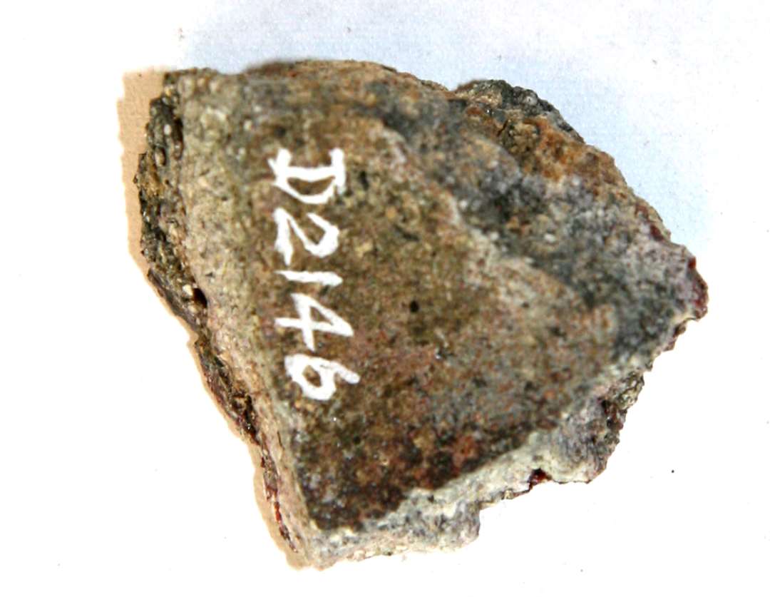 1 side- eller bundsk�r fra smeltedigel af gr�ligt, por�st, sandblandet gods, der p� ydersiden er d�kket af en sintret, uj�vn, lerkappe, hvis overflade fremtr�der som et r�dgr�t, glasuragtigt lag.