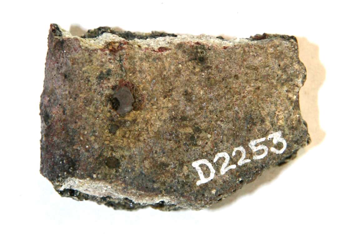 1 mundingsrandsk�r fra smeltedigel af gr�ligt, por�st, sandblandet gods. Ydersiden er d�kket af en uj�vn sintret skorpe, hvis overflade fremtr�der som et gr�ligt, glasuragtigt lag. Under skorpen anes en glat mundingsrand med indadskr�nende overkant.