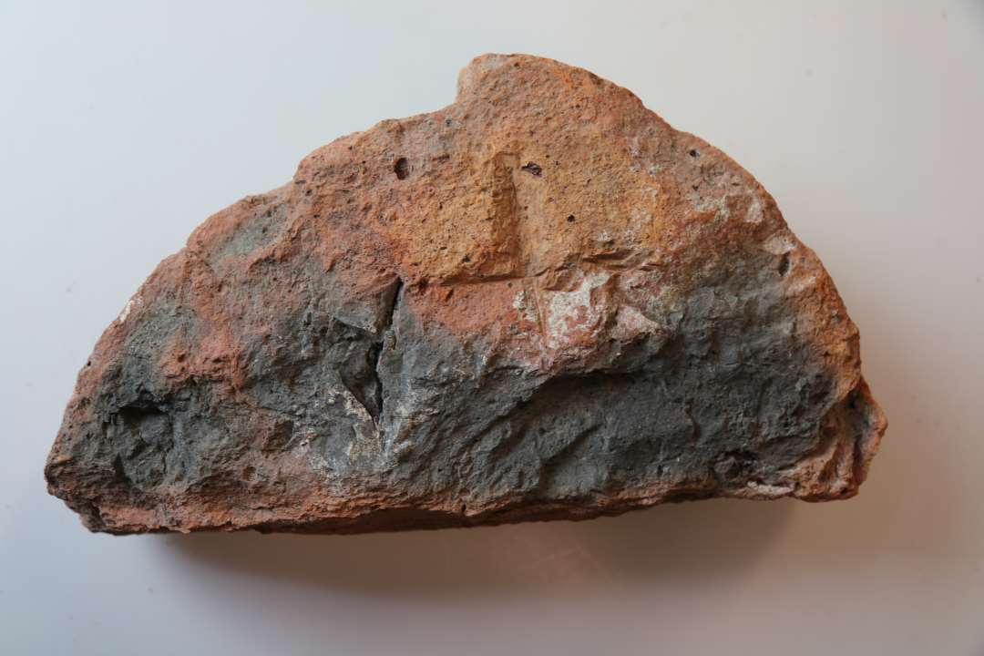 En halvrund munkesten med et indhak p� rundsiden. Der er indhakket et kors/grafitti p� undersiden.�M�l ca. 24 x 13 x 7 cm. 