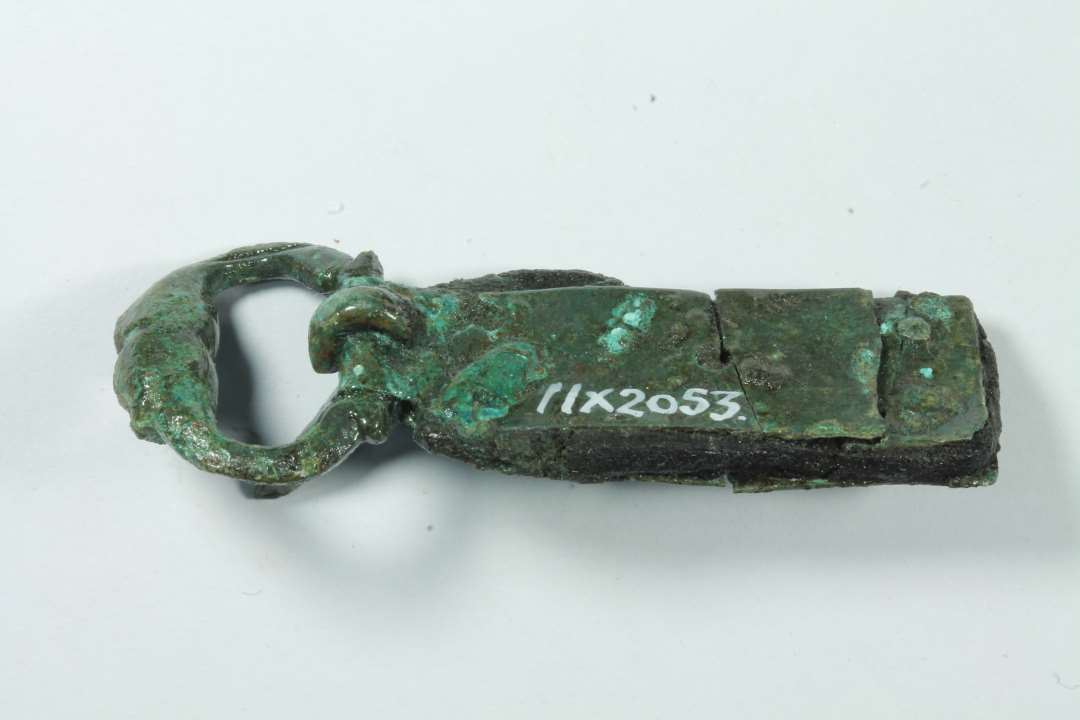 1 stk sp�nde og 1 fragment evt. af samme.��Fint, lille D-formet st�bt sp�nde med de kobberblikplader bevaret, som har holdt selve remmen fast. P� pladerne punslet ornamentik. Rest af l�derrem bevaret mellem nittepladerne. Genstandens samlede l�ngde: 5,2 cm., Rembeslagets bredde: 1,4 cm.Sp�nderammens st�rste m�l: ca 2,1 cm.