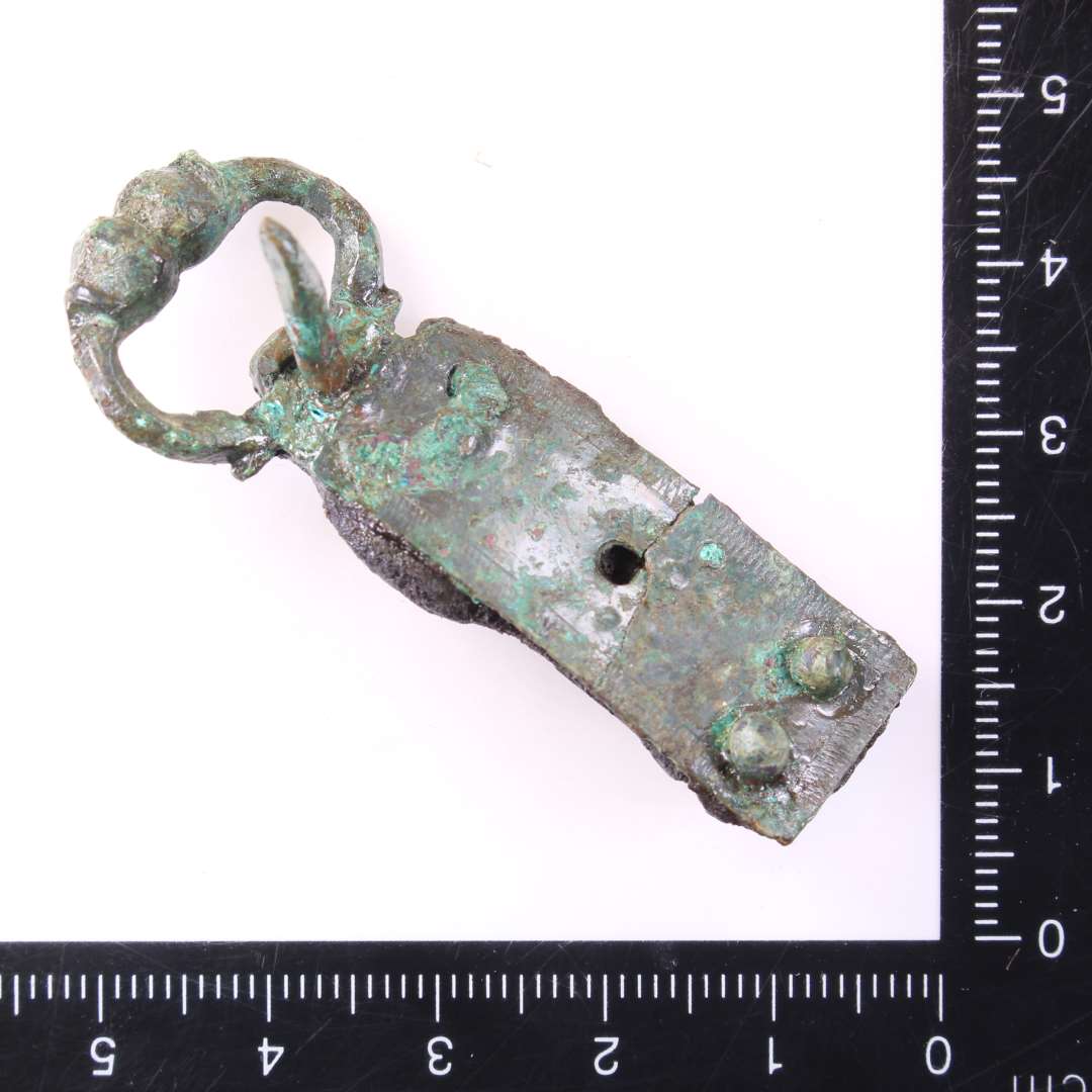 1 stk sp�nde og 1 fragment evt. af samme.��Fint, lille D-formet st�bt sp�nde med de kobberblikplader bevaret, som har holdt selve remmen fast. P� pladerne punslet ornamentik. Rest af l�derrem bevaret mellem nittepladerne. Genstandens samlede l�ngde: 5,2 cm., Rembeslagets bredde: 1,4 cm.Sp�nderammens st�rste m�l: ca 2,1 cm.