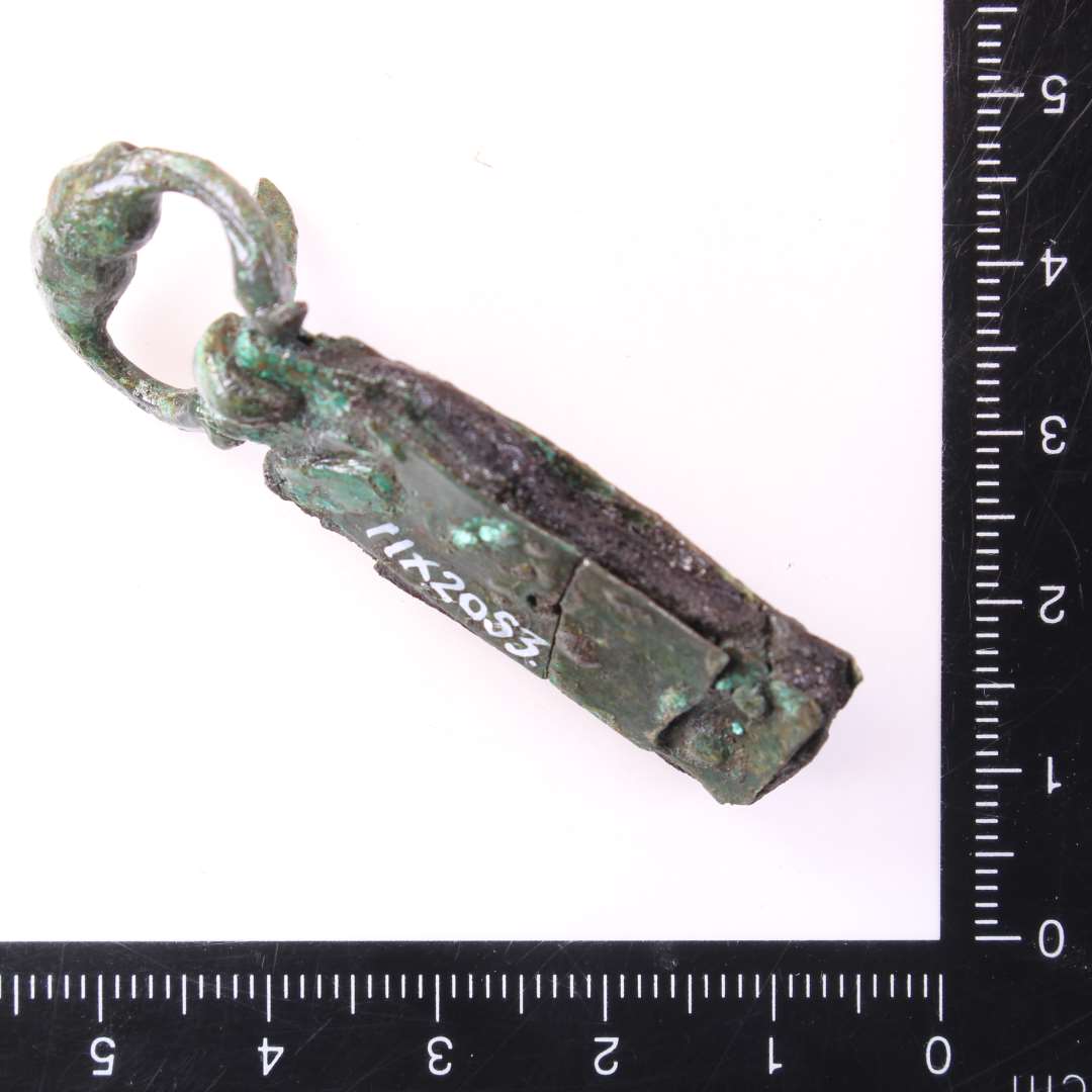 1 stk sp�nde og 1 fragment evt. af samme.��Fint, lille D-formet st�bt sp�nde med de kobberblikplader bevaret, som har holdt selve remmen fast. P� pladerne punslet ornamentik. Rest af l�derrem bevaret mellem nittepladerne. Genstandens samlede l�ngde: 5,2 cm., Rembeslagets bredde: 1,4 cm.Sp�nderammens st�rste m�l: ca 2,1 cm.