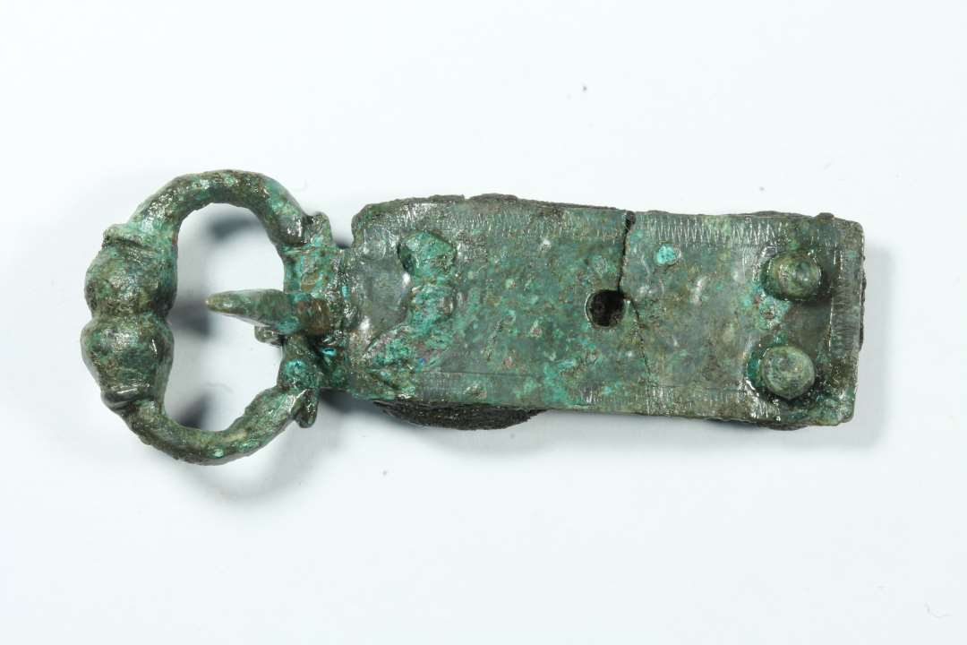 1 stk sp�nde og 1 fragment evt. af samme.��Fint, lille D-formet st�bt sp�nde med de kobberblikplader bevaret, som har holdt selve remmen fast. P� pladerne punslet ornamentik. Rest af l�derrem bevaret mellem nittepladerne. Genstandens samlede l�ngde: 5,2 cm., Rembeslagets bredde: 1,4 cm.Sp�nderammens st�rste m�l: ca 2,1 cm.