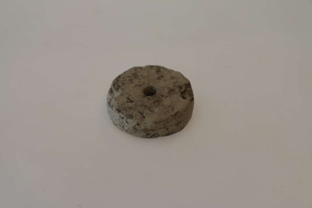 Skiveformet genstand med hul i midten. Kalk/kridtsten. 2 cm. tyk, 6 cm. i diameter med hul p� 0,8 cm. i midten��Anvendelse uvis, for let som v�vev�gt, for tung som tenv�gt, selv om den sidste anvendelse ikke kan udelukkes.