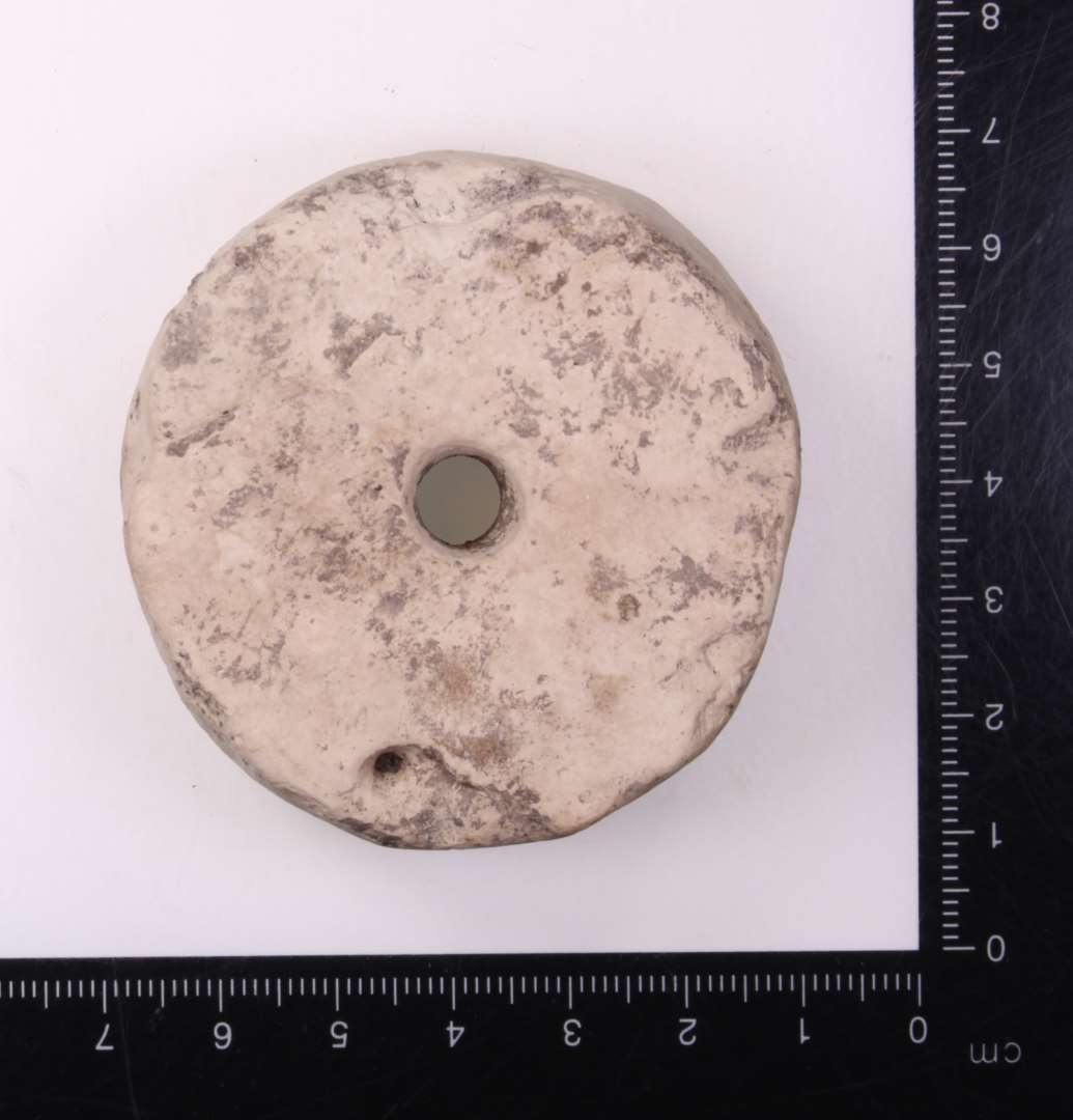 Skiveformet genstand med hul i midten. Kalk/kridtsten. 2 cm. tyk, 6 cm. i diameter med hul p� 0,8 cm. i midten��Anvendelse uvis, for let som v�vev�gt, for tung som tenv�gt, selv om den sidste anvendelse ikke kan udelukkes.
