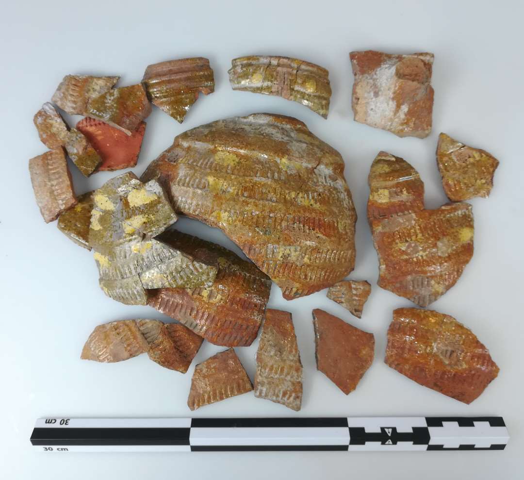 Fra samme kande. Best�r ogs� af sk�r fra x247. Flamsk (?) rullestemplet rundbuget kande p� b�lgefod, � 13 cm. Opr. h�jde ca. 25 cm. Udsmykket med tilf�ldigt udseende pletter af pibeler p� korpus og skulder. Bugen er helt d�kket af oml�bende simple rullestempler. Profileret, let fortykket rand. hvor der ogs� synes at have v�ret dekoration med pibeler og p�lagt lerdekoration., som m�ske har dannet et hoved (dyr/menneske?) 