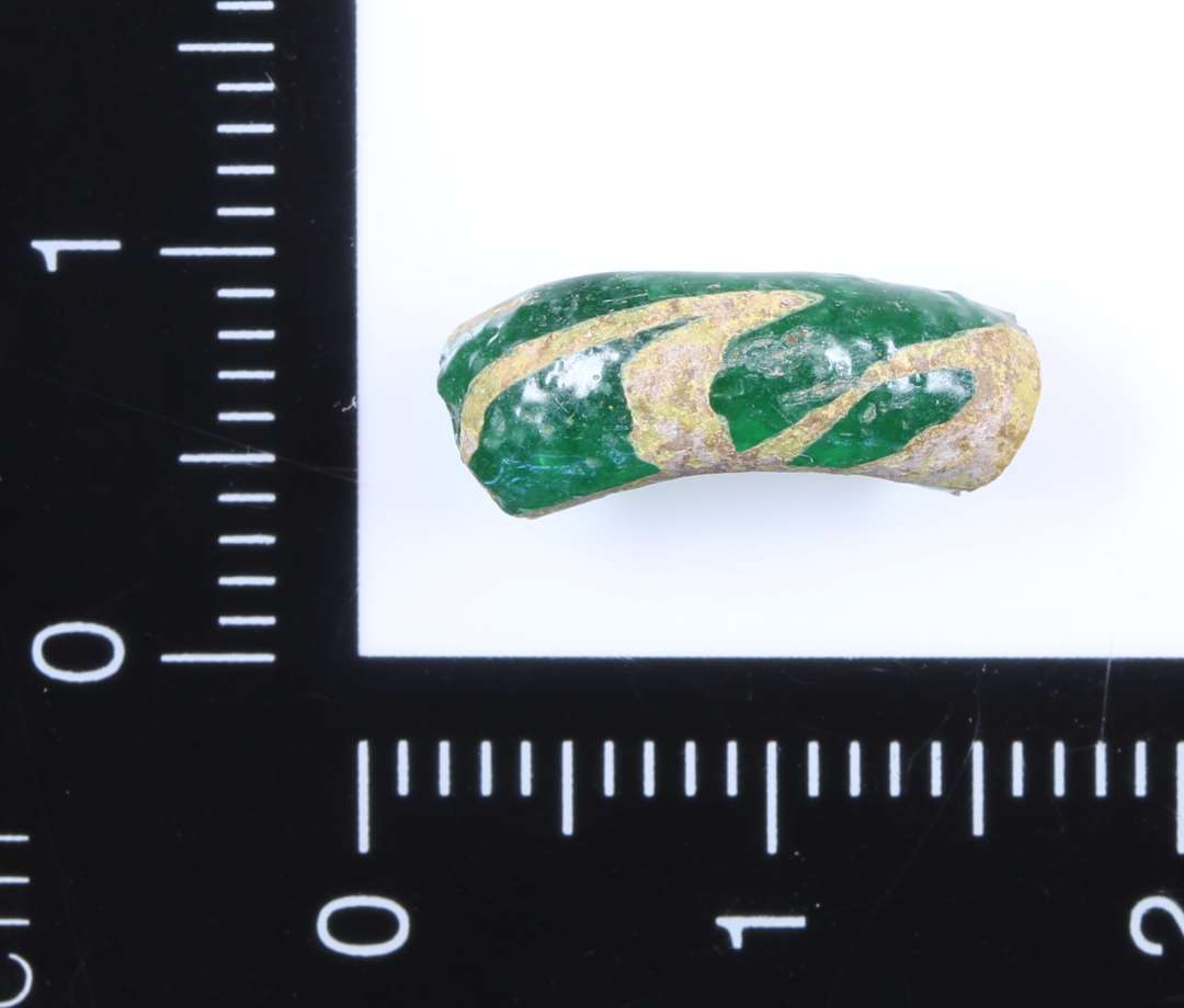 St�rre fragment af glasring i klart gr�nt glas. P� ydersiden er indlagt b�lgede streger af mat hvidgult materiale. Materialet er kun indlagt p� overfalden. Formodet diameter p� ca. 2 cm.