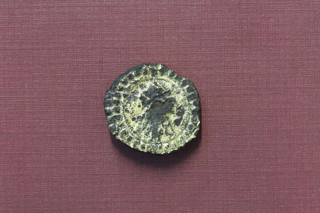 Karolingisk pseudo-m�ntfibula. Noget medtaget p� overfladen, der ellers generelt er med bevaret forgyldning. Der anes en h�jrevendt buste med ansigt i profil, antagelig med dragt og evt. med diadem eller (str�le?-)krone. Midterfladen er mod kanten afgr�nset af inderst en ganske fin og tyndperlek�de og yderst af 1-2 r�kker grove perler. P� bagsiden ses rester af n�lef�ste og -skede. St�rste diameter: 26 mm.