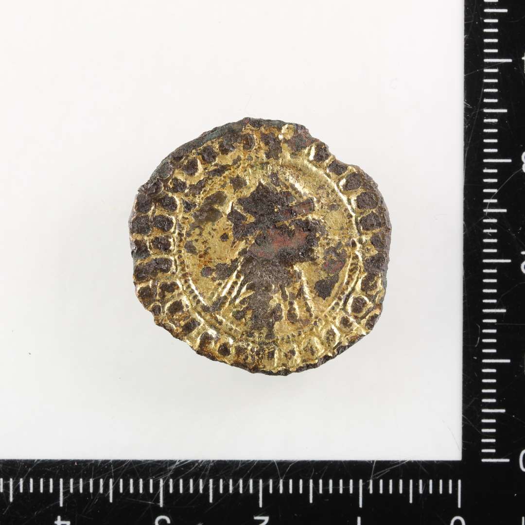 Karolingisk pseudo-m�ntfibula. Noget medtaget p� overfladen, der ellers generelt er med bevaret forgyldning. Der anes en h�jrevendt buste med ansigt i profil, antagelig med dragt og evt. med diadem eller (str�le?-)krone. Midterfladen er mod kanten afgr�nset af inderst en ganske fin og tyndperlek�de og yderst af 1-2 r�kker grove perler. P� bagsiden ses rester af n�lef�ste og -skede. St�rste diameter: 26 mm.