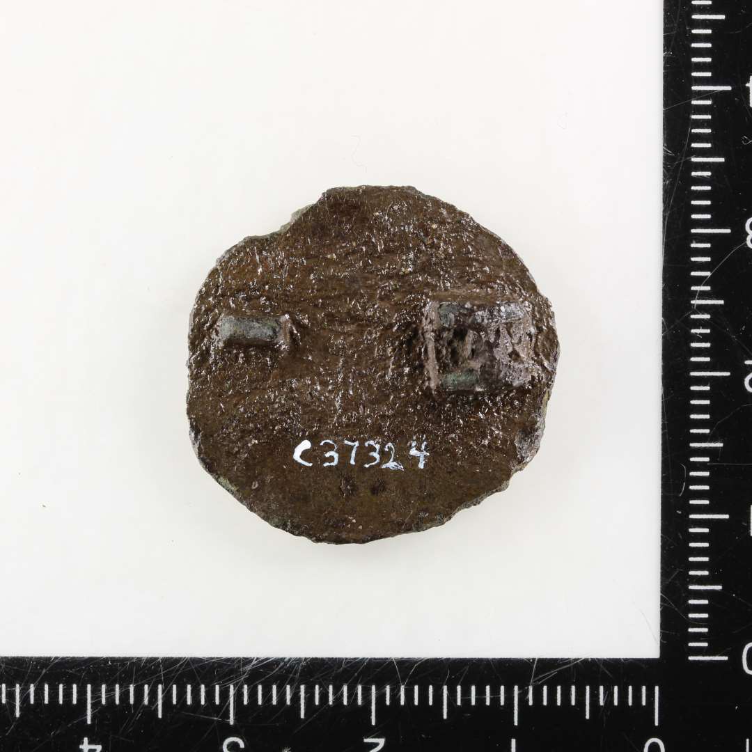 Karolingisk pseudo-m�ntfibula. Noget medtaget p� overfladen, der ellers generelt er med bevaret forgyldning. Der anes en h�jrevendt buste med ansigt i profil, antagelig med dragt og evt. med diadem eller (str�le?-)krone. Midterfladen er mod kanten afgr�nset af inderst en ganske fin og tyndperlek�de og yderst af 1-2 r�kker grove perler. P� bagsiden ses rester af n�lef�ste og -skede. St�rste diameter: 26 mm.