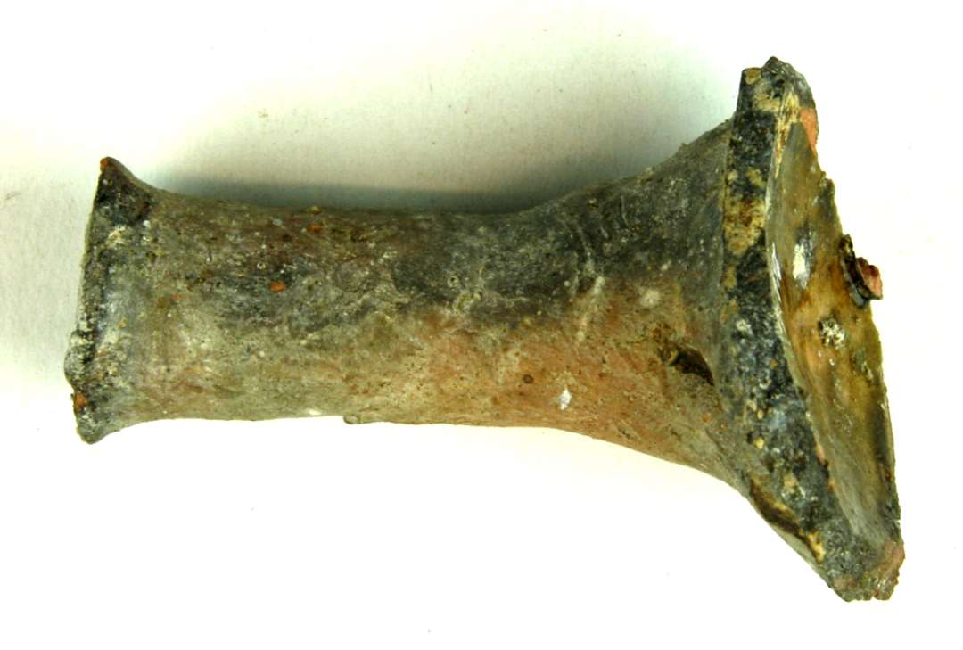 Stjerthank af hul, glat r�rform, let udvidet og lige oversk�ret i stjertenden. Stykket er st�rkt sintret, oprindelig af r�dbr�ndt lergods, men fremtr�der nu gr�br�ndt. Indersiden er d�kket af klar blyglasur.