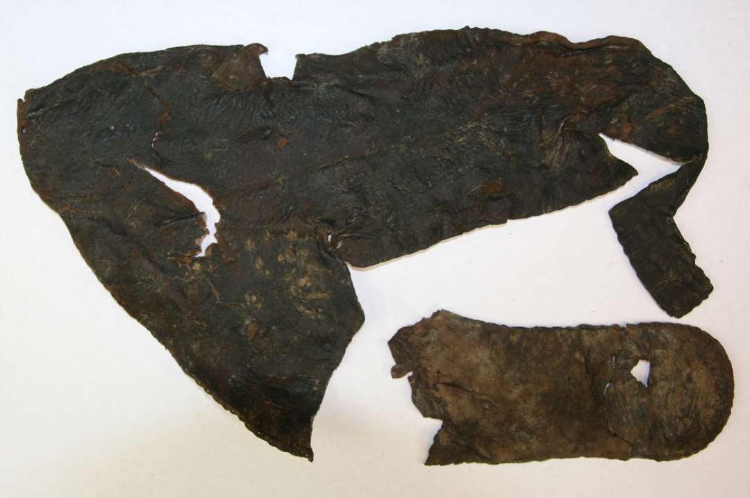 Fragment af overl�der af sko til halvvoksen person, antagelig fra h�jre fod. Overl�deret har bevaret de syede rande langs s�lkanten og fod�bning. Overl�deret er omtrent helsk�ret og dets samling er foreg�et i venstre side langs en stumpvinklet tilsk�ring, str�kkende sig fra midten af vristen ned under ankelknoen, hvor der i kanten mod s�len har v�ret indf�jet en ubetydelig (nu manglende)kile. Fod�bningen afsluttes midt p� vristen med et par opst�ende ca. 2,5 cm. h�je flipper med ca. 1,5 cm. dybe vanrette indsk�ringer ved dets rod. Skoen har haft markeret n�sespids. Spor af nogle lukke- eller sn�reanordninger forefindes ikke. Partiet af fod�bningen i venstre side ned mod h�len er bortsk�ret. L�ngde fra h�l til t�: ca. 25,0 cm. Sideh�jde udfor ankel: ca. 8,0 cm. (total h�jde: 10 cm.)