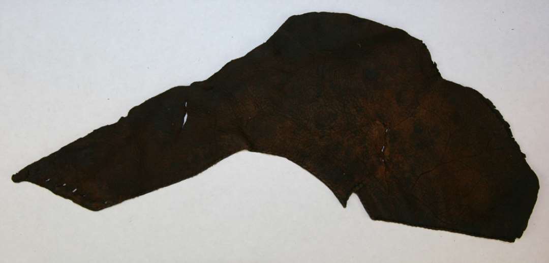 Fragmenteret overl�der af sko, antagelig til h�jre fod hos halvvoksen person. Alle overl�derets rande mod s�len er afrevet samt hele t�partiet. En st�rre skr�kile i venstre side mangler. Fod�bningen er lige oversk�ret i vristen, men s�ledes at der p� hver side af vristryggen har v�ret en lille fremstikkende, spids flip, hvoraf kun den ene er bevaret, idet den anden har udgjort en del af den manglende kiles overkant. Fod�bningens rand viser syspor efter et nu manglende kanteb�nd. Skoen er dybt nedsk�ret i ankelsiderne og i den venstre side er overl�deret skr�t tilsk�ret og forsynet med 7 t�tsiddende sn�rehuller, idet de 7 tilsvarende huller har v�ret placeret i den nu manglende kile i nedsk�ringens modsatte side. P� indersiden af overl�deret er der under sn�rehullernes spor efter fastsyning af en nu manglende ca. 2 cm. bred forst�rkende l�derstrimmel. Overl�derets bevarede h�jde udfor vrist: ca. 5,0 cm. Overl�derets bevarede h�jde udfor h�l: ca. 6,0 cm. Overl�derets st�rste l�ngde: ca. 35,0 cm.