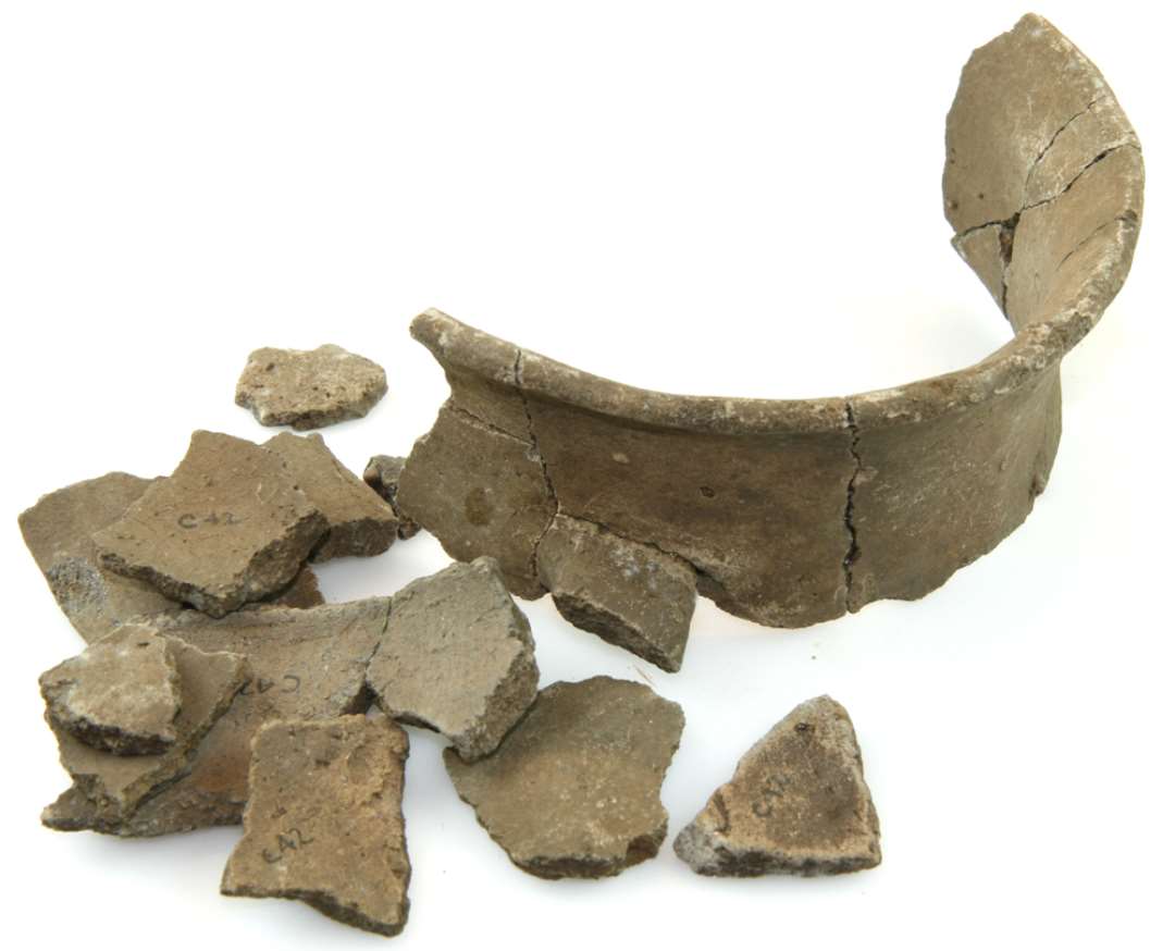 Sk�r fra lerkar, enhanket med flad bund, konvexe sider og afsat konkav hals og lige afsk�ret udfaldende rand, 1 lodret b�ndformet �re fra halsens nederste del til ? skulder. P� skulderen ornamentik af dobbeltfurer i zigzag af smalle dobbeltfurer i vinkel i de trekanter, der fremkom mellem zigzaglinierne. Brunt, j�vnt, glat gods, delvist st�rkt br�ndt sekund�rt. Ca. � delen af halsen og sk�r af bug og rand bevaret. Randdiameter: ca. 15 cm.�