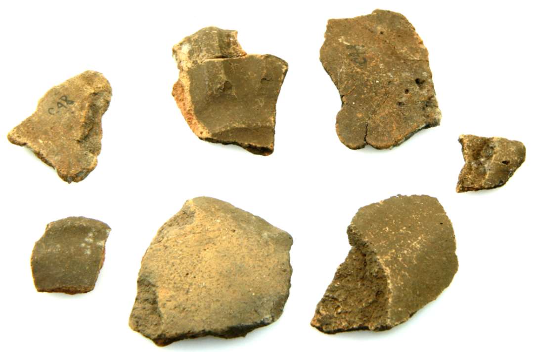 Sk�r af lerkar med flad bund, konvekse sider. P� skulderen 1 lodret aflang knop med brede lodrette furer p� begge sider. Ogs� skr� modstillede furer p� skulderen. P� halsen vandrette furer. Brunt, glat, fint gods stedvist r�dbr�ndt sekund�rt. Tykkelse: 0,4 cm.