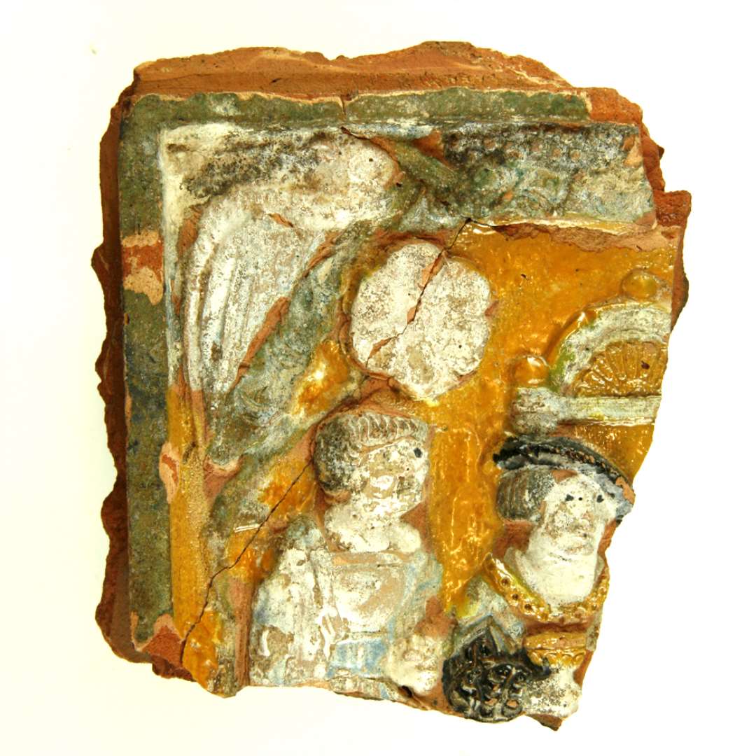 Fragment af polykrom kakkel. Motiver er lignelsen om den rige mand og Lazarus. �verste venstre hj�rne. Sammenlimet af tre stykker. I midten mand med pageh�r og m�rkebrun baret, med gylden k�de om halsen og gylden b�ger i h�nden, h�jre. Ovenover arkitektur (fra tronstol). Til venstre mand med kort brunt h�r, oversk�g og bl� kl�dning. Langs kanterne bladv�rk, og i hj�rnerne papeg�je. Indenfor hvid blomst. Uprofileret ramme. Sammenlimet med D1619. St�rste m�l: 14 cm.