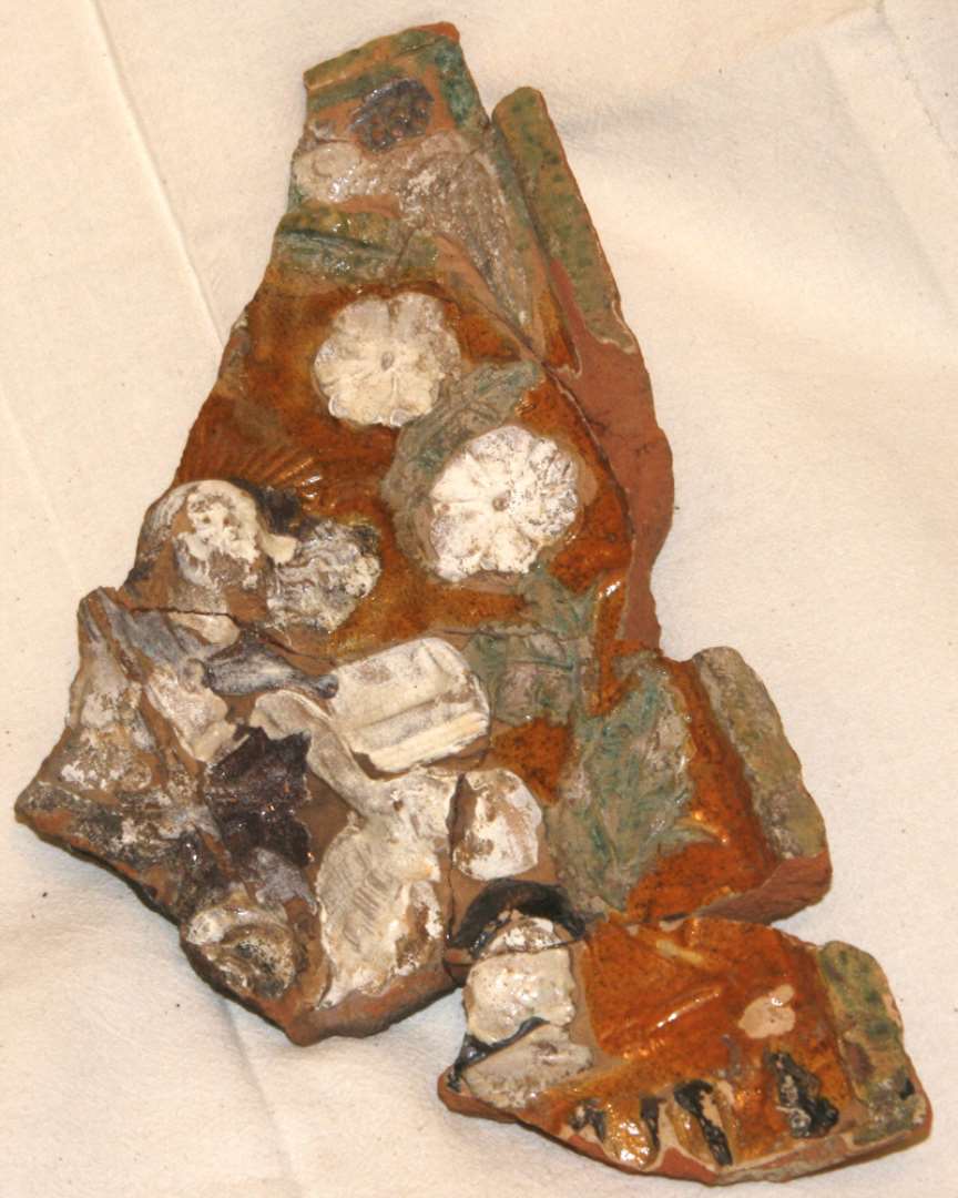 Fragment af polykrom kakkel. �vre venstre trediedel mangler. Sammenlimet af 8 stykker. I midten Kristusfigur med str�leglorie, brunt h�r og sk�g, med l�s brun kappe, med stav (korsfane?) i h�jre h�nd og med sin venstre st�ttende p� hovedet af liggende skelet. Bag hem en kiste. Han st�r p� liggende mandsfigur med lang brun kappe og brun hue. Langs rammen bugtede st�ngler med gr�nne blade og hvide blomster. I hj�rnet pappeg�je. P� et postament forneden indskriften: 'ann (0-1) ii -' med gotiske bogstaver, brune. Uprofileret ramme.�Oprindelig bredde: 18,8 cm. L�ngde: 25,8 cm.