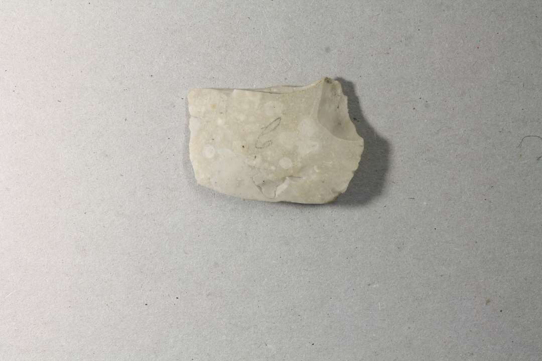 �ksefragment, lys, mat, sp�ttet flint; afslag, hvis slagflade udg�res af den ene �gflade og dorsalflade af den anden �gflade, af en sleben �kse. 3,1 x 4,3 x 0,8 cm.