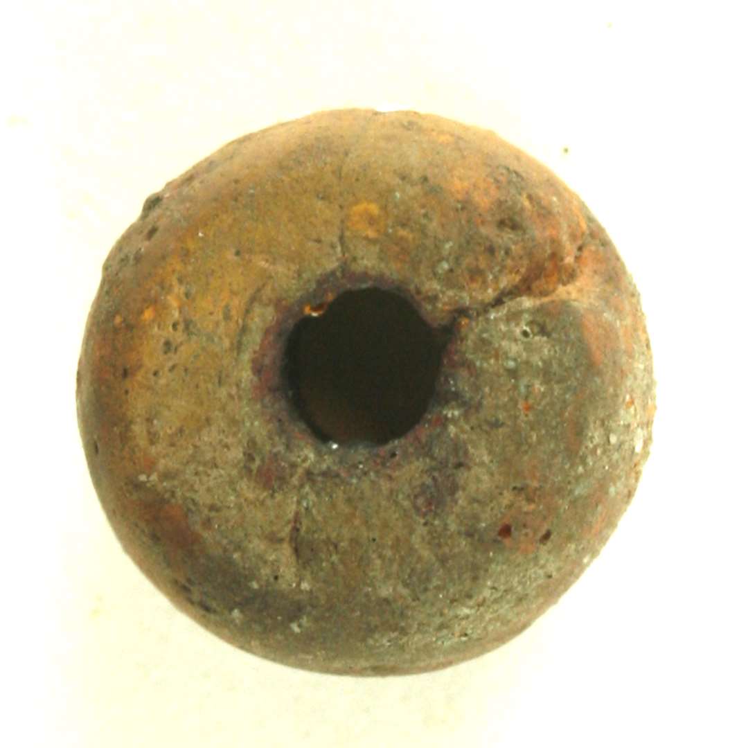 Perle af br�ndt ler i gr�brun, r�dbrun og brunlige farvetoner. Perlen har let hv�lvet t�ndeform og er lige afsk�ret i begge ender, hvor gennemboringernes mundinger findes lidt sk�vt anbragt. en ca. 2,5 cm. lang, uj�vn fordybning findes i kanten af den hv�lvede side. St�rste bredde: ca. 1 cm. H�jde: ca. 0,75 cm. Gennemboringens diameter: ca. 0,3 cm.