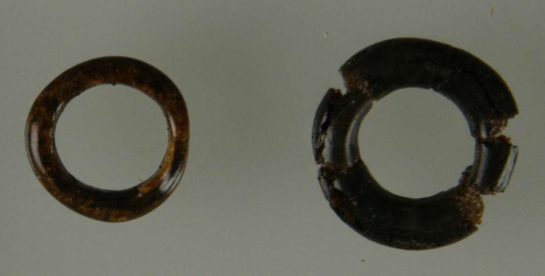 To glasringe. Den ene, sammenlimet af fire stykker, er af nu mat, gr�sort, uigennemsigtigt glas, 1,8-1,85 cm i diam., udf�rt af en ca 0,45 cm tyk p�lseformet glastr�d, der p� ringens sammenf�jningssted kun andrager godt 0,3 cm. Den anden af st�rkt uregelm�ssig cirkelform er af blankt gyldenbrunt nu uigennemsigtigt glas, fra 1,5-1,6 cm i diam. Ringp�lsens tykkelse varierer fra knap 0,3 cm til 0,2 cm p� sammenf�jningsstedet.