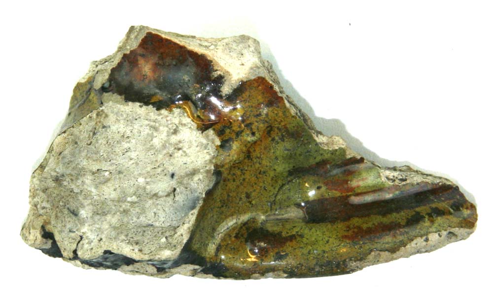 Randsk�r antagelig af kande med afbrudt hank. Gr�lig h�rdtbr�ndt pibelersagtigt gods, p� ydersiden forsynet med partiel begitning af r�dler, dels under l�beranden, dels under hankens basisparti, alt d�kket af klar til gr�nlig blyglasur. Indersiden d�kket af gr�nligsp�ttet blyglasur. Fragmentet udviser kraftig l�berand med plan overflade, hvorunder spor af svage omkringl�bende furer og vulster. Hanken, der antagelig har v�ret af trind p�lseform, viser p� oversiden spor af to kraftige furer med en svagere midt imellem.