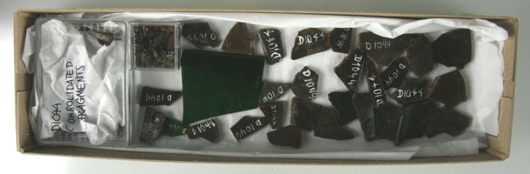 Den bevarede sk�rpr�ve (ca 50 stykker) best�r af sortbrune, uigennemsigtige, ildsk�rnede (?) fragmenter p� n�r to sm�stumper (ca 0,8 og 1 x 1,7 cm) og et st�rre stykke (ca 4,5 x 5 cm) med afrundet fortykket sidekant, der alle tre har bevaret en klar konsistens og dybgr�n farve.�Selvom enkelte af sk�rene danner ret krumme flader er de fleste dog temmelig plane med en tykkelse varierende fra 1,5 - 4 mm. Ti af sk�rene udviser en glat afrundet fortykket sidekant (indtil 5 mm tyk), hvorimod ingen viser spor efter afnapning i kanterne.