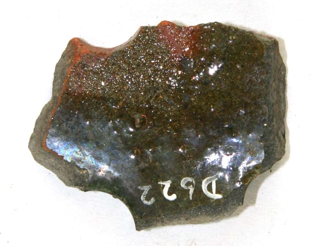 Bundsk�r, 1,0 - 1,4 cm. tykt, af gr�br�ndt lergods. I sk�rets kanter findes tre tydelige og et svagt spor af cirkul�re huller boret ret sk�vt igennem sk�rv�ggen f�r br�ndingen. B�de sk�rets over- og undersiden samt hullernes sidev�gge er d�kket af gr�nlig blyglasur. 