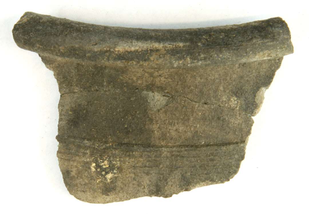 1 mundingsrandsk�r af gr�br�ndt fintmagret lergods fra kar med fortykket, afrundet udadb�jet rand med hulkehl p� indersiden. P� skulderen findes en flad afsats med t�tte omkringl�bende furer og vulster. Gr. 9.