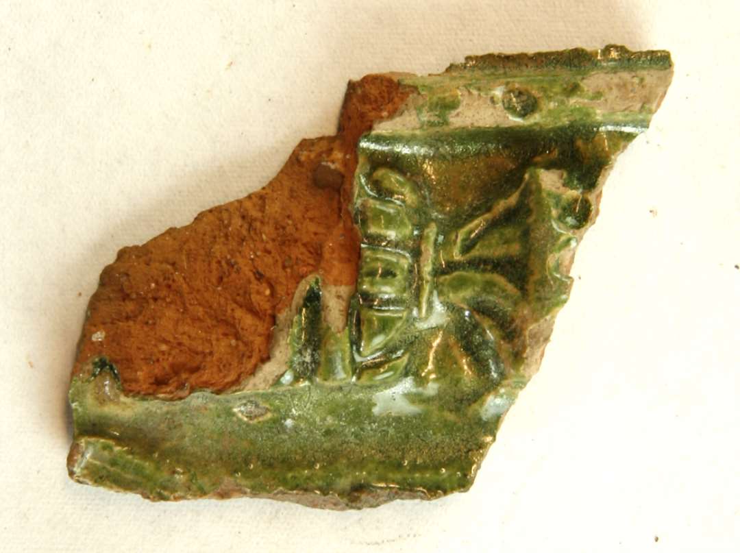 1 fragmentstump af gr�nglaseret kakkel med rest af stiliseret ornamentik p� ydersiden. St�rste m�l: 5,5 cm.