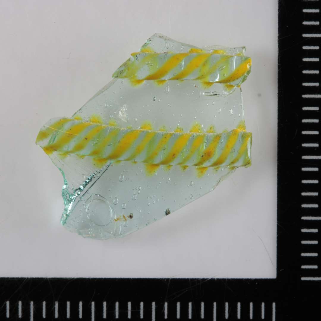 1 sidesk�r fra kar (b�ger) af gennemsigtigt svagt gr�nligt glas, der p� ydersiden er belagt med to kraftige vulstformede tr�de af samme masse, hvilke er forsynede med p�smeltede tynde flade b�nd af ugennemsigtig gullig masse i tovsnoet forl�b. Reticella.