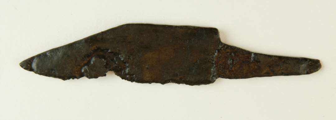 Knivsblad af jern med flad skafttunge, hvis underside flugter med bladets �g, men er fors�nket i forhold til bladryggen. Bladet har en langstrakt spids, der danner en stump vinkel i forhold til bladryggen. L: 9 cm, heraf skafttynge 3 cm. Br: 2 cm.