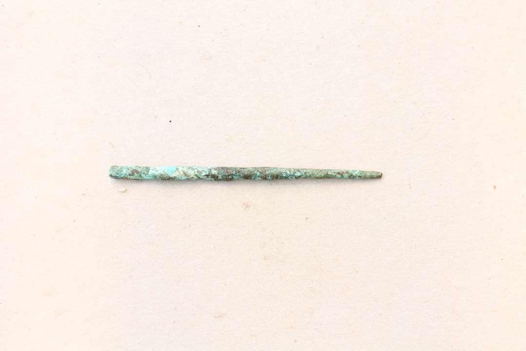 1 noget forvitret n�l af bronze af trind ganske svagt b�jet form.�L: 6,6 cm. Diameter i den tykke ende: 0,35 cm. Ikke noget �je at se, muligvis afbr�kket.