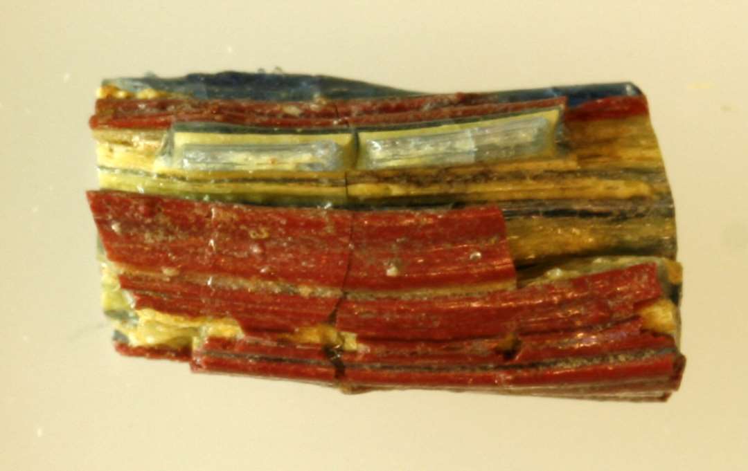 1 stykke af svagt b�jet glasstang af noget sk�v, kvadratisk tv�snitsform samt en samling fragmenter af samme. Stangen er dannet af sammensmeltede tr�de af gennemsigtigt bl�t og ugennemsigtigt gulligt glas, der i tv�rsnit fremtr�der i et skaktavlagtigt m�nster. Overfladen er delvis d�kket af et sammensmeltet lag af tr�de af r�dbrun ugennemsigtig glasmasse.