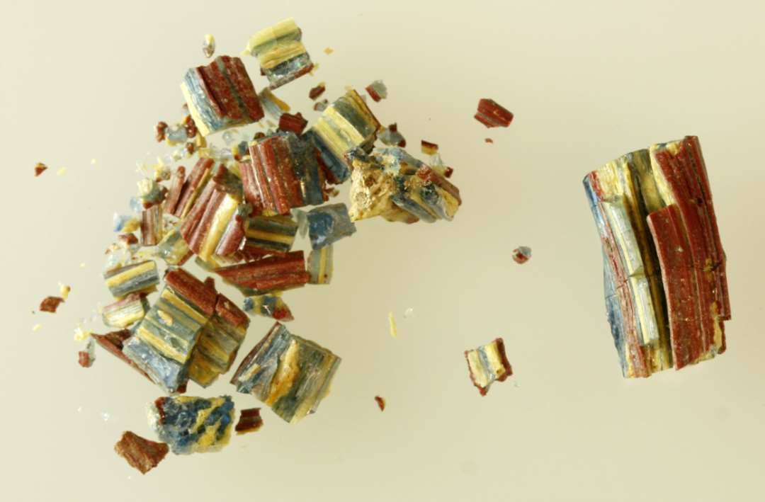 1 stykke af svagt b�jet glasstang af noget sk�v, kvadratisk tv�snitsform samt en samling fragmenter af samme. Stangen er dannet af sammensmeltede tr�de af gennemsigtigt bl�t og ugennemsigtigt gulligt glas, der i tv�rsnit fremtr�der i et skaktavlagtigt m�nster. Overfladen er delvis d�kket af et sammensmeltet lag af tr�de af r�dbrun ugennemsigtig glasmasse.