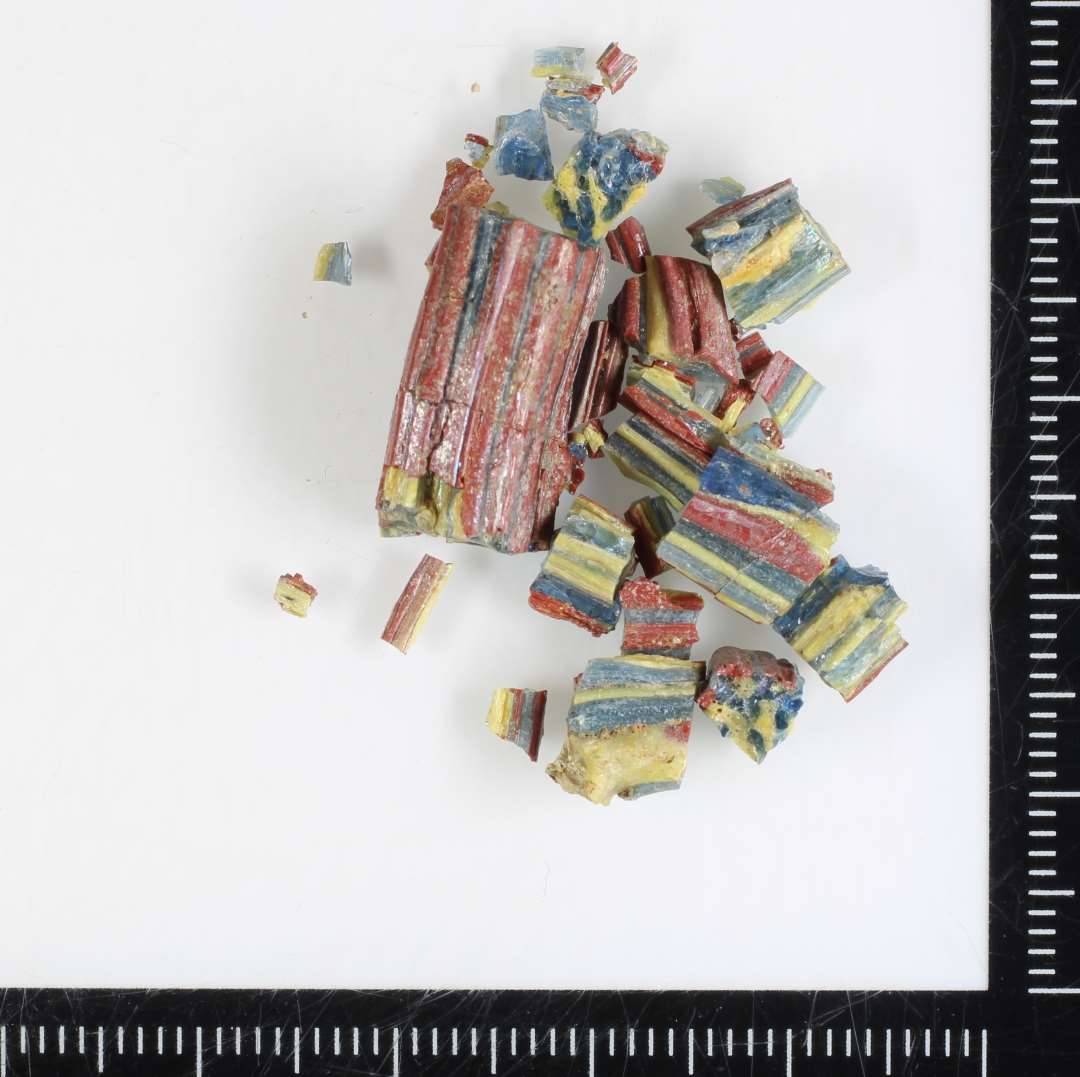 1 stykke af svagt b�jet glasstang af noget sk�v, kvadratisk tv�snitsform samt en samling fragmenter af samme. Stangen er dannet af sammensmeltede tr�de af gennemsigtigt bl�t og ugennemsigtigt gulligt glas, der i tv�rsnit fremtr�der i et skaktavlagtigt m�nster. Overfladen er delvis d�kket af et sammensmeltet lag af tr�de af r�dbrun ugennemsigtig glasmasse.