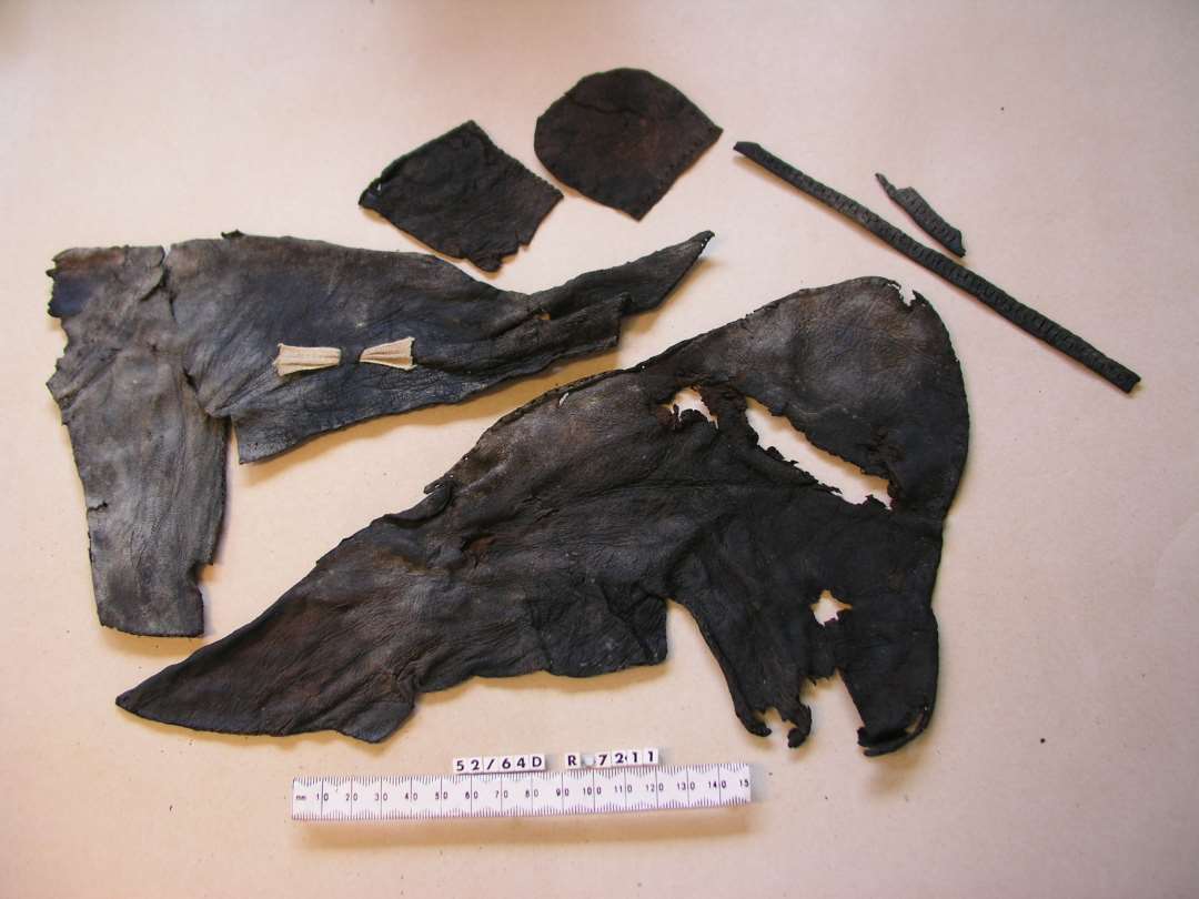 11 stk. l�der, heraf 6 stk.bevaret: St�rkt indt�rret og forrevet Overl�derfragment af sko antagelig til halvvoksen Person. Nedenfor Udsk�ringen der danner Fodaabningens ene Forkant findes et lille tv�rgaaende oprindelig sammensyet Indsnit. Sammensyning ved Skraasnit i Siden hvor en kile der har dannet en Del af Fodaabningen og dens anden Forkant mangler. S�mspor af Kantbaand langs den bevarede Del af Fodaabningens Rand. Ingen Spor af Lukkeanordning. �St�rste Maal ca 19 x 33 cm. St�rkt forrevet og indt�rret Overl�derfragment af Sko antagelig til halvvoksen Person. Nedenfor Udsk�ringen der danner Fodaabningens ene Forkant findes et lille tv�rgaaende Indsnit. Skraa Tilsk�ringssnit i Siden, hvor en Kile, der har dannet en Del af fodaabningen og dennes anden Forkant mangler. S�mspor langs den bevarede Rand af fodaabningen fra et kantbaand. Midt p� Siden ca 2,5 cm. under fodaabningens Kant findes et Par lodrette gennemstukne �jer, som en Sn�rerem har l�bet igennem. St�rste Maal ca 18 x 28 cm.�Kantbaand, l�der: Fragment af Kantbaand antagelig fra Sko. Tildannet af en ca 1,5 cm. bred L�derstrimmel, der er omb�jet og forsynet med lodrette gennemskaarne �jer anbragt med 2 � 3 mm's Mellemrum. Tydelige S�mspor langs Randene paa Underkanten, hvor der n�r Stykkets ene Ende findes et bueformet Indhak af Hensyn til Baandets Omb�jning. St�rste L�ngde ca. 22,5 cm. St�rste Bredde ca. 0,7 - 9,8 cm. Saalfragment, l�der: Separat tilskaaret H�lparti af Saal af tyndt L�der (Bindsaal) til voksen Person. St�rste Maal ca. 6 x 7 cm. Overl�derfragment (?): Lille L�derstykke af sk�v rektangul�r Form med s�mspor langs to af Kanterne N�r Stykkets ene Hj�rne samt omtrent paa dets Midte findes et gennemskaaret �je. St�rste Maal ca. 6 x 7 cm.