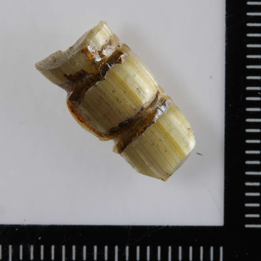1 fragment af glasperle. Aflang cylindrisk. Stribet gr�nligt uigennemsigtigt glas med tv�rstriber af m�rkere farve. ( Sektioneret ).