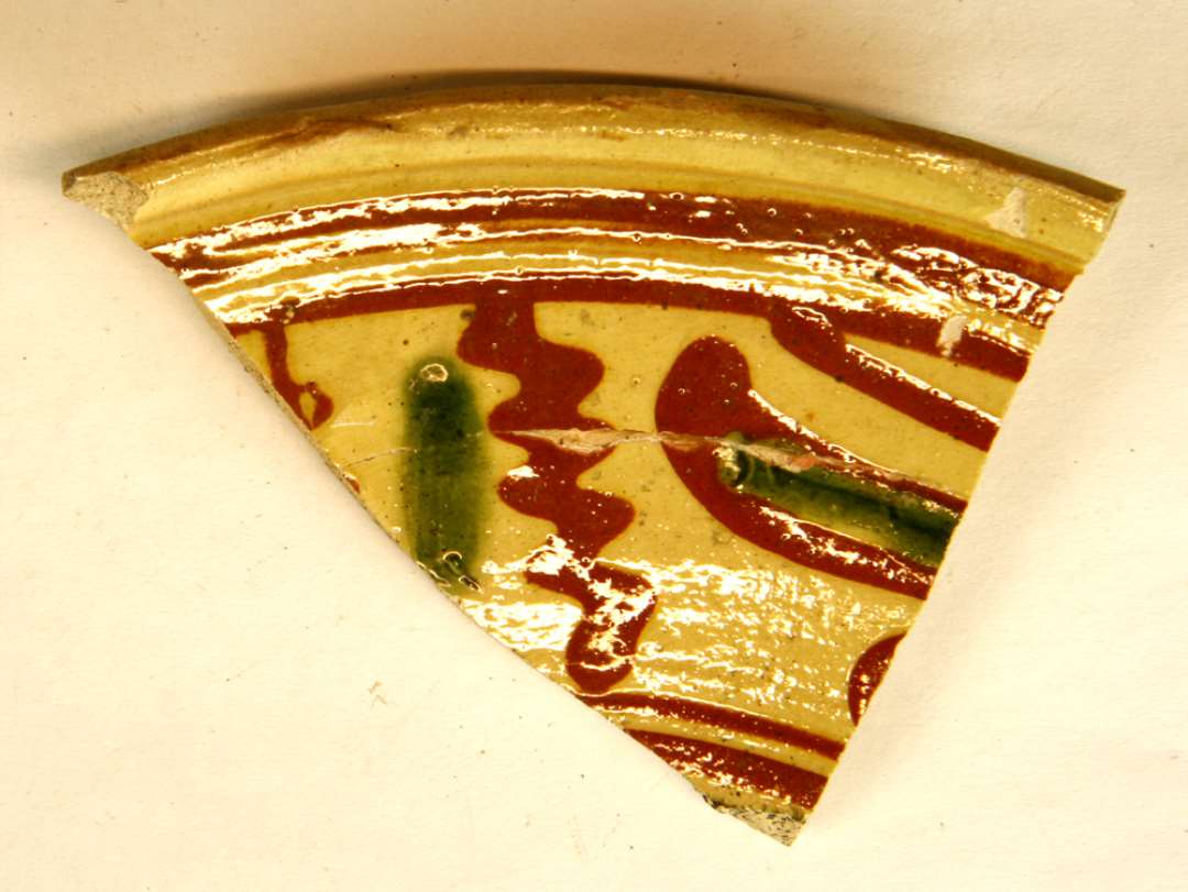 1 randsk�r af hornmalet fad af gulligt lergods med pibelersbegitning og dekorationer i r�dler og gr�nt p� indersiden, hvorover klar blyglasur, gruppe 1.�