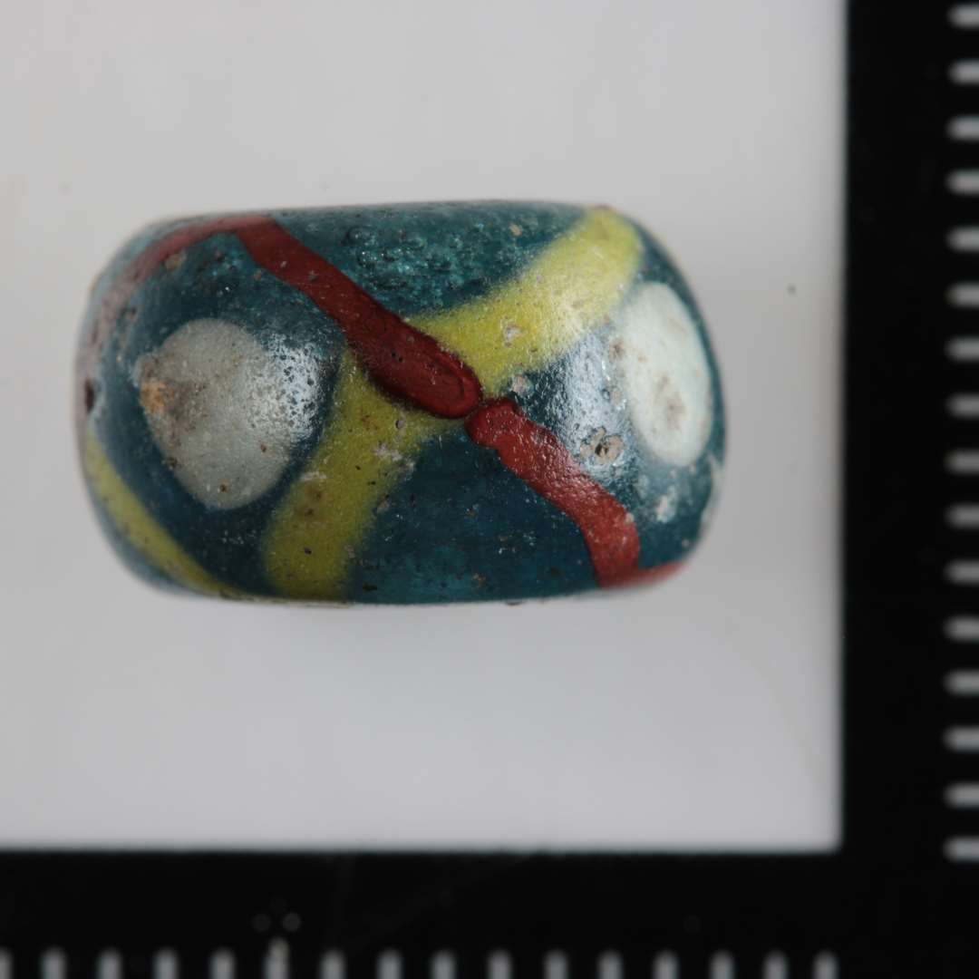 1 fragmenteret skiveformig afrundet perle af gennemsigtigt bl�ligt glas med korsvis indlagte gule og r�dbrune tr�de i kanten afvekslende med cirkul�re indl�gninger af hvidt glas.