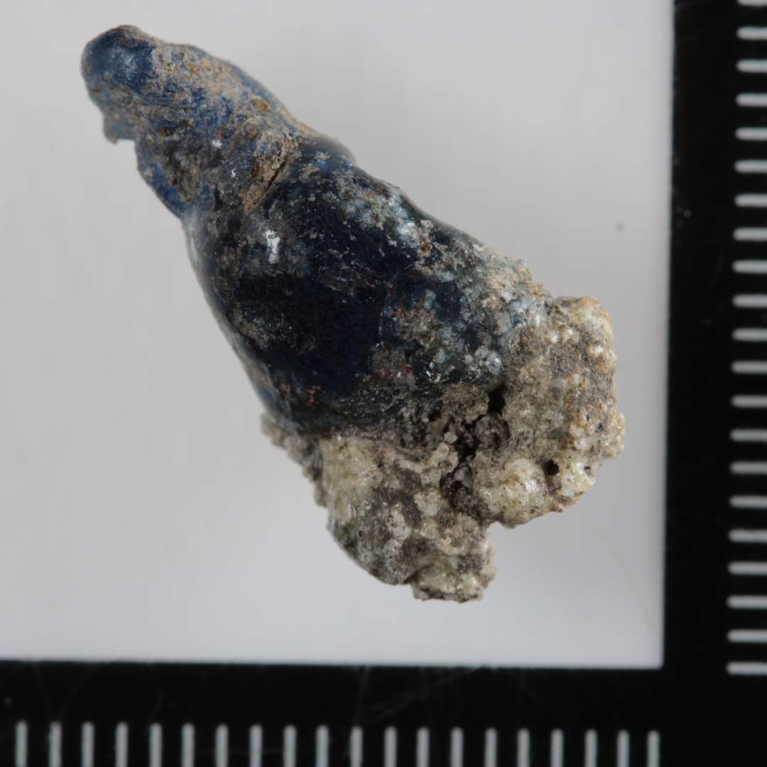 1 kr�mmerhusformet stump smeltet glas af bl�ligt gennemsigtig glasmasse i hvis brede ende er fastsmeltet en stump sintret lerslagge.�L�ngde: 1,9 cm.