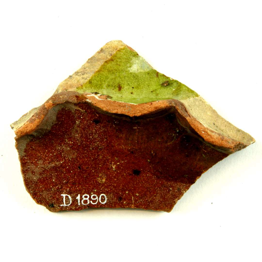 Bundsk�r af r�dbr�ndt lergods fra flamsk kande med fremst�ende b�lgende fodrand dannet ved kraftige fingerindtryk. P� ydersiden er sk�ret forsynet med pibelersbegitning, delvis d�kket af gr�nlig blyglasur. Bunden er p� ydersiden og indersiden d�kket pletvis af klar blyglasur.