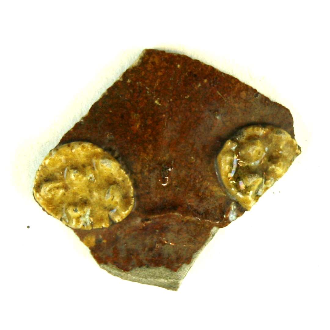 Bugsk�r af gr�ligt por�s pibelersagtigt gods fra kar med bred flad halsfure. P� ydersiden er lagt en tynd r�dlersbegitning ovenp� hvilken er anbragt to delvis bevarede knoprosetter. Hele ydersiden er d�kket af klar blyglasur, s�ledes at rosetterne fremtr�der gullige p� en r�dbrun baggrund.