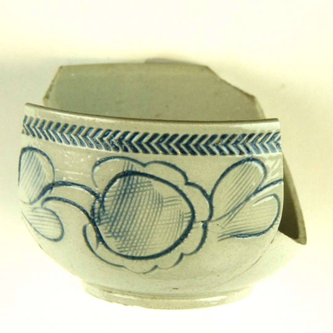 2/3 af en kop af gr�t stent�j, samlet af to sk�r. Cirkul�r standkant, halvkugleformet korpus med blomster og blade i indridset relief p� siden og under mundingen indpresset Zig-zagfrise. I dekorationens bund bl� farve. Bunddiameter: 3,4 cm. H�jde: 4,0 cm. Mundingsrand: 7,5 cm.