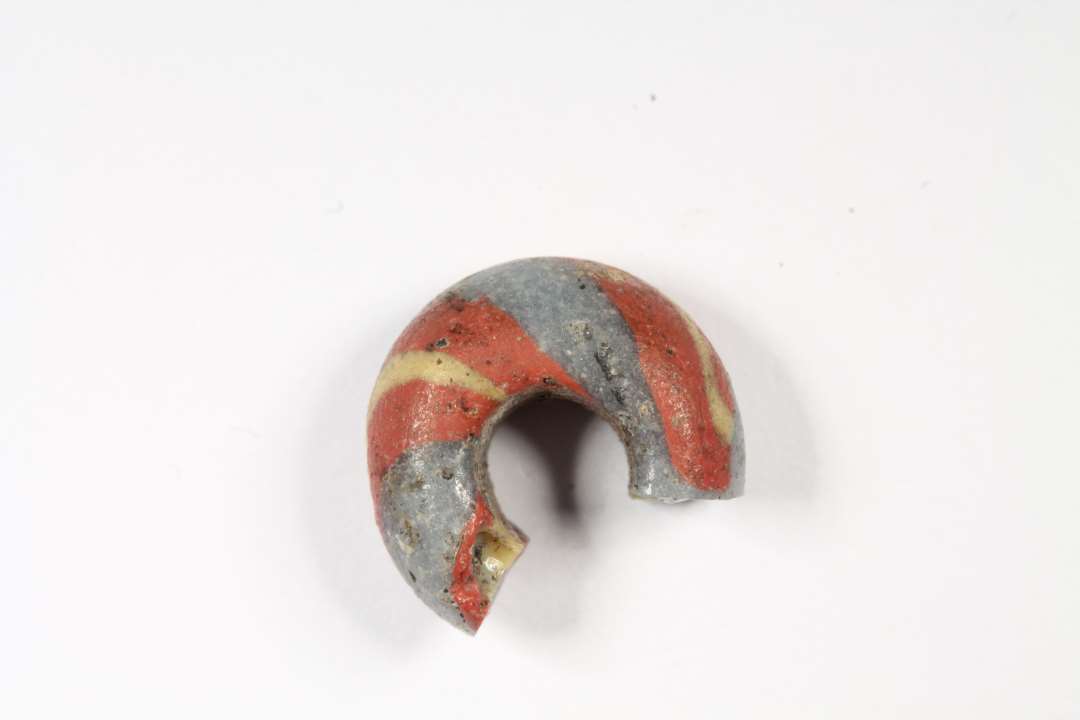 Fragment af ringformet, uigennemsigtig lysebl� glasperle med r�de og gule striber. Diameter: 1,1 cm.