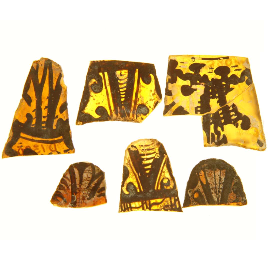 6 fragmenter af ruder af gult glas med afnappede kanter, bemalet med stiliserede plantemotiver i brunlig farve. Hidr�rer antagelig fra ruder af afsmalnende tungeform med afrundet spids og svag konkav indb�jning ved roden. De 4 af fragmenterne hidr�rer fra tungespidsen, 2 fra tungeroden. St�rste bredde og l�ngde: 2,15 x 1,9 cm. - 2,5 x 1,9 cm. - 3,3 x 2,6 cm. - 2,45 x 2,8 cm. - 2,8 x 4,0 cm. - 3,9 x 3,7 cm.
