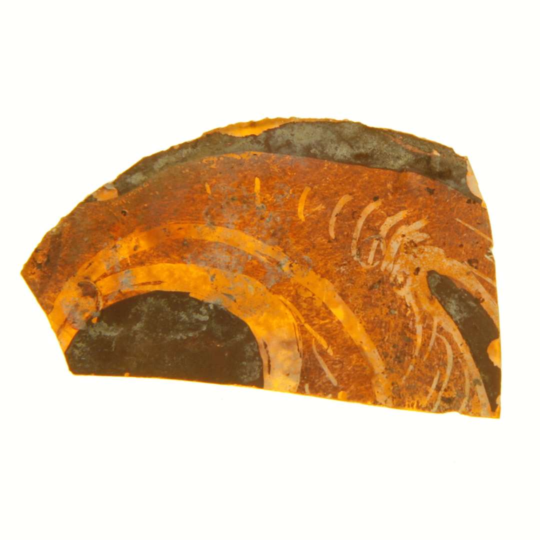 1 fragment af rude af gult glas med afnappede kanter bemaling med spiralslyngninger, der fremtr�der klare og gennemsigtige p� en baggrund af brunlige farver, dels tynde og n�sten gennemsigtige dels p�f�rt i tykke ugennemsigtige lag. Hidr�rer antagelig fra rude af cirkul�r form. St�rste m�l: 2,8 x 4,8 cm.