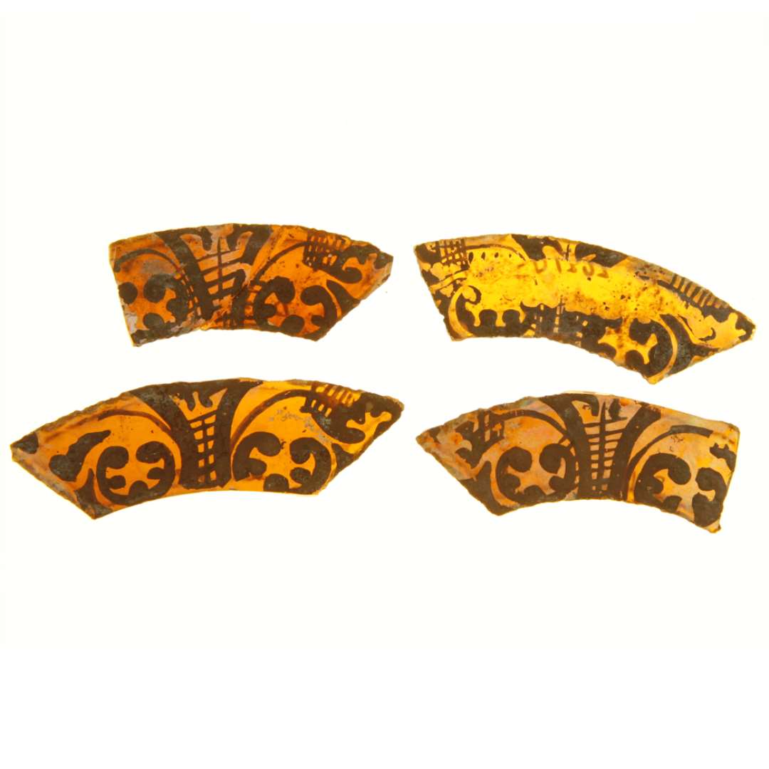 4 omtrent hele ruder af gult glas med afnappede kanter, alle bem�lede med omtrent samme stiliserede plantemotiv i fraftige p�f�rt brun farve. Ruderne har krum rektangul�r form, s�ledes at langsiderne forl�ber paralelt i krum linief�ring, medens enderne er skr�t afsk�ret. Man kunne kalde dem b�ndformede. 2 af ruderne mangler endehj�rne. St�rst l�ngde og bredde: 5,2 x 2,3 cm. - 5,9 x 2,4 cm. - 6,6 x 2,35 cm. - 7,3 x 2,4 cm.