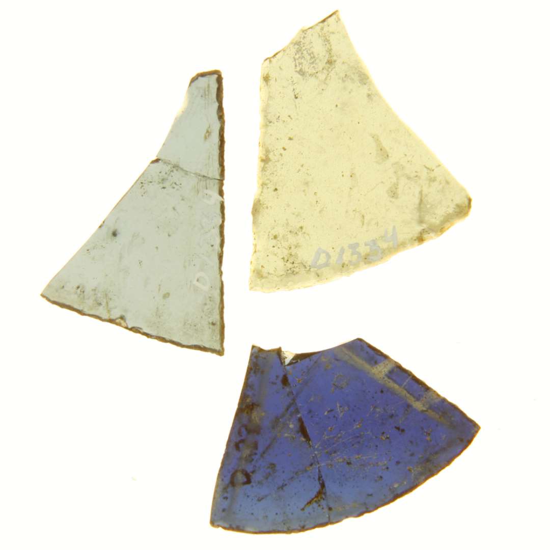 3 rudefragmenter af bl�ligt glas tildels med afnappede kanter hidr�rende fra ruder med form som et cirkeludsnit med en centrumsvinkel p� ca. 65 grader. 2 af fragmenterne der hver er sammenlimet af 2 stykker, udm�rker sig ved en s�rdeles kraftig dybbl� farve. St�rste m�l: 4,2 x 3 cm. - 4,3 x 2,9 cm. - 3,75 x 3,6 cm.